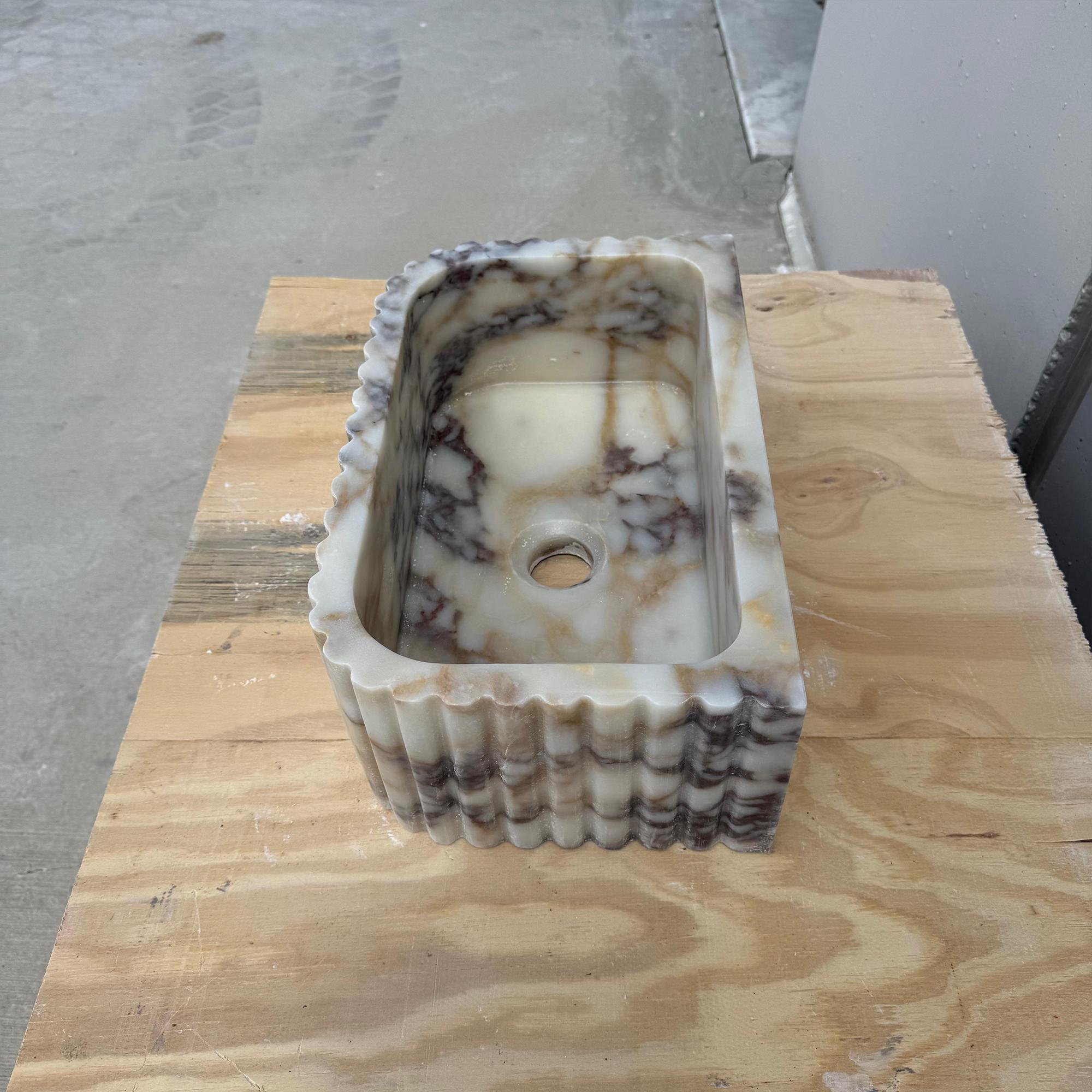 A rectangular marble sink with detailed fluting rests on a rustic wood countertop.