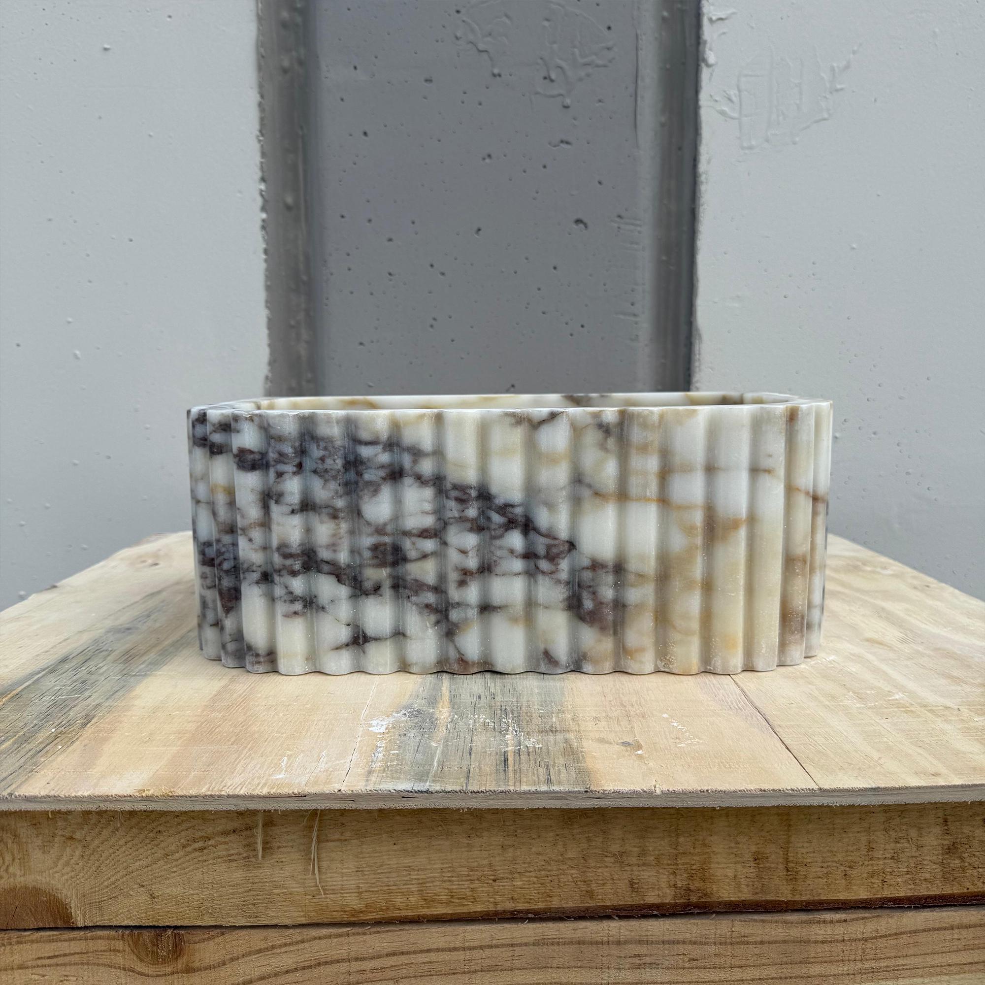 A small marble vessel sink with fluted edges is displayed in a minimal studio setting.