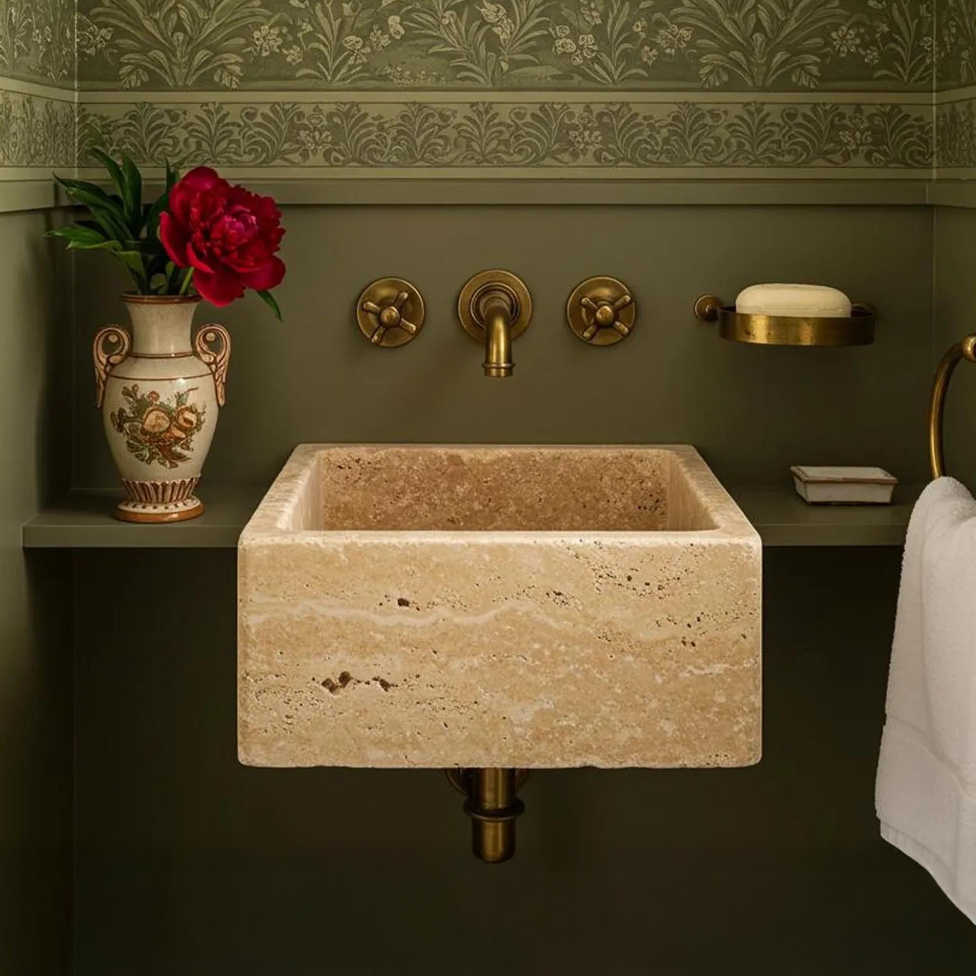 A wall-mounted rectangular travertine sink featured against a deep olive green backdrop with brass fixtures.